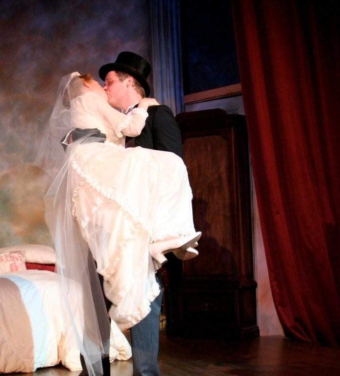 Actor Kevin Curran (Michael) carries Margaret Smith (Agnes) across the threshold in The Montgomery Theatre production of The Fourposter.  The bridal dress is a reproduction of the one worn by L.M. Montgomery for her own wedding in 1910.